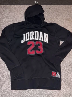 Jordan svart hoodie med tryck - Säljer en svart hoodie från Jordan med stort vitt JORDAN-tryck och röd 23 på bröstet. Klassisk huva och ribbade muddar. Ikonisk patch med Jumpman-logga och nummer 23 nertill. Perfekt för dig som gillar sportig streetstyle. XS 