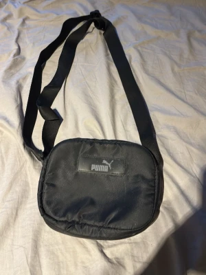 Svart axelväska från Puma - Snygg svart axelväska från Puma med justerbar rem och diskret logga på framsidan. Väskan har en mjuk, vadderad känsla och är tillverkad i slitstarkt syntetmaterial. Perfekt storlek för det viktigaste och enkel att bära crossbody. Lite smutsig men går o tvätta bort de