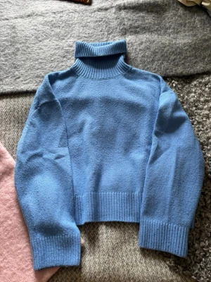 Blå polotröja i stickat material - Mysig blå stickad polotröja med bred ribbstickad kant vid ärmslut, nederkant och krage. Tröjan har en rak, något boxig passform och är tillverkad i ett mjukt material som känns skönt mot huden. Perfekt för kyliga dagar och enkel att styla.
