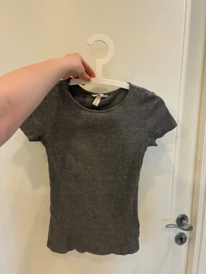 Grå ribbad t-shirt från H&M - En enkel och stilren grå ribbad t-shirt från H&M med rund halsringning och korta ärmar. T-shirten har en figurnära passform och är tillverkad i ett mjukt, stretchigt material som sitter snyggt på kroppen.