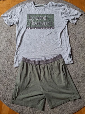 Grå pyjamas från Bruno Banani XL - Snygg pyjamas från Bruno Banani i storlek XL. T-shirt i ljusgrått med stort tryck framtill och texten 'not for everybody'. Matchande shorts i olivgrönt med elastisk midja och logga. Perfekt för chillkvällar hemma.