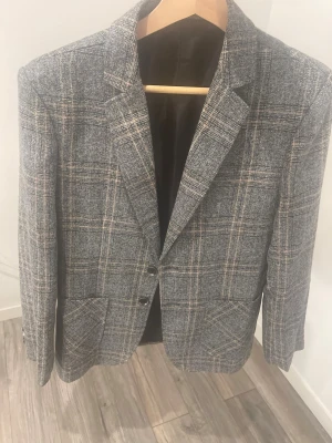 Grå rutig kavaj i ullmix - Snygg grå kavaj med rutigt mönster och två knappar framtill. Klassisk passform och mjukt material, perfekt för en stilren look. Kavajen har två fickor och är helfodrad. Passar till både skjorta och t-shirt.