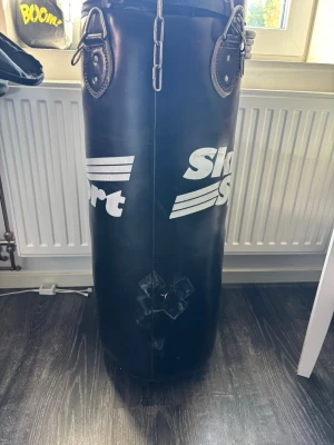 Svart boxsäck från Sport Sport - Boxsäck i svart konstläder från Sport Sport med vita logotyper på sidan. Säckens form är cylindrisk och den har metallkedjor för upphängning. Perfekt för träning hemma eller på gymmet.
