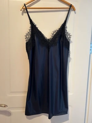 Svart satin nattlinne med spets - Elegant svart nattlinne i satin med tunna axelband och vacker spetsdetalj längs v-ringad hals. Materialet är glansigt och mjukt, vilket ger en lyxig känsla. Perfekt för dig som vill ha något stilrent och bekvämt att sova i.