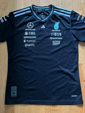 Mercedes F1 Team T-shirt 2024 - Adidas (Storlek M) - Skick: Bra skick (använt i 6 manader) Storlek: M (Medium) - Herr/Unisex Märke: Adidas x Mercedes-AMG Petronas F1 Team Saljer en Mercedes-AMG Petronas Formula One Team driver t-shirt från säsongen 2024 Anänd i ungefär 6 manader men fortfarande i fint skick! Finns självklart lite spår av tvätt och användning, priset är inte hugget i sten.
