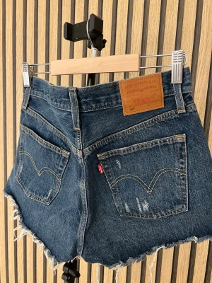Levi's blå jeansshorts med slitningar - Säljer ett par klassiska blå jeansshorts från Levi's med rå kant och slitna detaljer framtill och baktill. Shortsen har fem fickor, hög midja och den ikoniska Levi's-lappen bak. Perfekta för sommaren och ger en avslappnad look. Använda en gång.