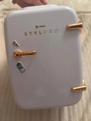 STYLPRO sminkkylskåp vit/guld - Snyggt och kompakt sminkkylskåp från STYLPRO i vit plast med guldfärgade detaljer. Kylskåpet har en liten hylla inuti och dekorerande stickers på utsidan. Perfekt för att förvara hudvård och sminkprodukter svalt och fräscht. Har haft den i bara några veckor