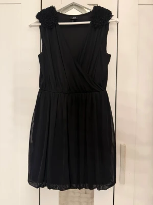 Svart omlottklänning från H&M -  En helt ny Svart kort omlottklänning från H&M med dekorativa blommiga axeldetaljer. Den är aldrig använd och finns inga fläckar eller något.  Klänningen är ärmlös, har v-ringning och ett lätt, flytande tyg som ger en snygg siluett. Perfekt för dig som vill ha en enkel men ändå unik look.