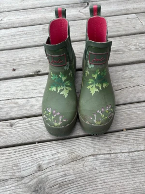Gröna blommiga gummistövlar Joules - Säljer ett par gröna gummistövlar från Joules med blommigt mönster och bladmotiv. Insidan är rosa och stövlarna har dragflikar bak för enkel påtagning. Perfekta för regniga dagar och trädgårdshäng.