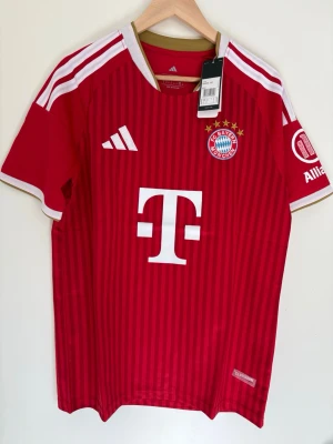Bayern München matchtröja Adidas M - Röd Bayern München matchtröja från Adidas med vita detaljer och klubbmärke på bröstet. Tröjan har guldfärgade inslag vid kragen, vita ränder på axlarna och stora sponsormärken framtill. Tillverkad i lätt och ventilerande material.