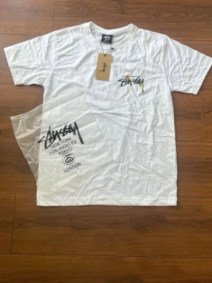 Vit Stüssy t-shirt med camo-logga - Vit t-shirt från Stüssy med deras ikoniska logga i camo-färger på bröstet. Klassisk passform och rund halsringning. Tillverkad i mjuk bomull, perfekt för en avslappnad streetwear-look.