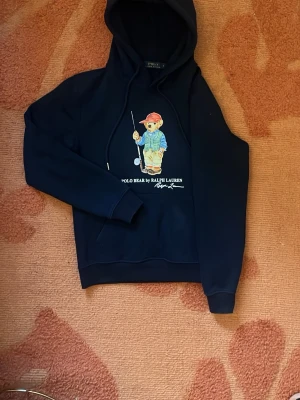 Svart hoodie Polo Bear Ralph Lauren - Snygg svart hoodie från Ralph Lauren med Polo Bear-tryck framtill. Motivet visar en björn med golfklubba och keps. Hoodien har huva med dragsko och ribbade muddar. Perfekt för dig som gillar streetwear och vill ha en ikonisk look.