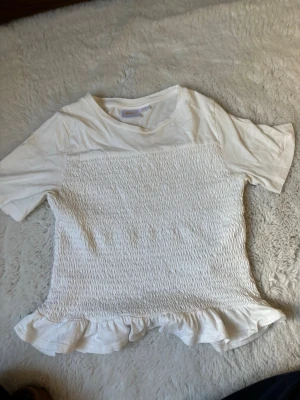 Vit topp med volang från Gina Tricot - Söt vit topp från Gina Tricot med korta ärmar och smockad front. Toppen har volangkant nedtill som ger en extra detalj. Perfekt för dig som gillar en enkel men ändå trendig stil.
