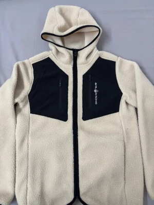 Vit teddyjacka från Sail Racing - Mysig vit teddyjacka med huva från Sail Racing. Jackan har svarta detaljer på bröstet med dragkedjeförsedda fickor och broderad logga. Tillverkad i fluffigt teddy-material och hel dragkedja framtill. Perfekt för kalla dagar och riktigt snygg streetstil. Original priset var på 1400kr. Helt oanvänd.