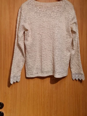 Crémefärgad spetsblus med strassdetaljer - Säljer en elegant vit spetsblus med långa ärmar och rund halsringning. Blusen har fina strassdetaljer framtill och vackra spetsmönster över hela plagget. Perfekt för dig som gillar romantisk och feminin stil. Stretchig och passar storlek L och uppåt. Perfekt till jobb, finare kvällar och även fester.
