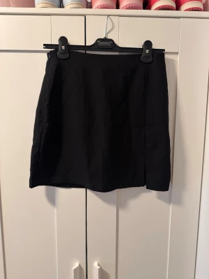 Svart kortkjol  - Enkel och stilren svart kortkjol med rak passform. Kjolen har en liten slits framtill för extra detalj och är perfekt att matcha med olika toppar. Klassisk design utan mönster eller synliga detaljer.