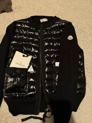 Svart cardiganjacka från Moncler - Svart cardiganjacka från Moncler med quiltad front i glansig polyester och stickade ärmar. Jackan har två fickor med knappar, dragkedja framtill och Moncler-logga på ärmen. Perfekt för dig som vill ha en stilren och exklusiv look.