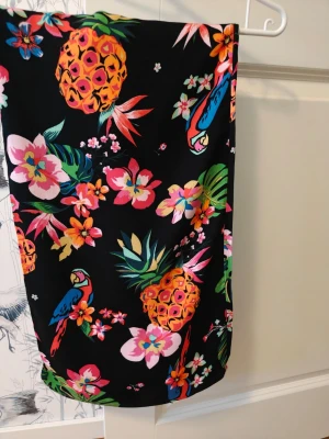 Färgglada byxor - Säljer ett par svarta byxor med tropiskt mönster av ananas, papegojor och blommor i starka färger som rosa, orange, blått och grönt. Midjan har en dragsko med tofsar. Perfekta för dig som vill sticka ut med en lekfull och somrig stil.