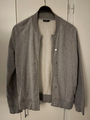 Grå hoodie med knappar från J.Lindeberg - Säljer en superfint och sparsamt använd grå bomberjacka i sweatshirt-material från det populära märket J.Lindeberg. Perfekt som ett skönt och stilrent lager-på-lager-plagg till våren, sommaren eller hösten.  