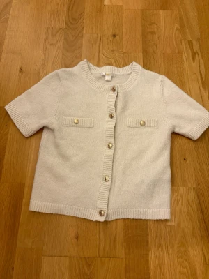 Kortärmad beige kofta med pärlknappar - Söt kortärmad kofta i beige med ribbade kanter och dekorativa pärlknappar framtill. Två fejkfickor med matchande knappar på bröstet ger en trendig look. Perfekt att styla över ett linne eller t-shirt.