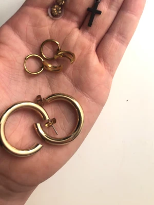 Mixade örhängen i guld och svart - Säljer ett set med flera örhängen: stora och små ringar i guldfärgad metall samt ett svart korsformat örhänge. Perfekt för dig som gillar att mixa och matcha olika stilar. Alla örhängen har enkel och trendig design.