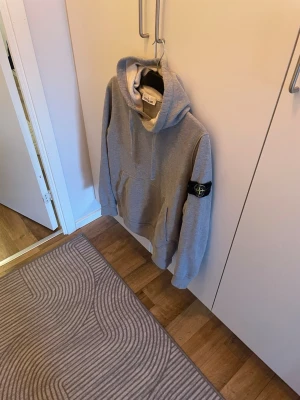 Grå hoodie från Stone Island - Snygg grå hoodie från Stone Island med klassisk patch på vänster ärm. Hoodien har en stor huva med dragsko och en rymlig magficka. Tillverkad i mjukt bomullsmaterial som är skönt att ha på sig. Riktigt finns skick 10/10 som ny kvitto finns