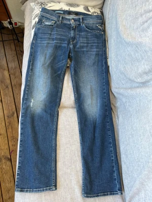 Blå jeans från Replay med raka ben - Säljer ett par blå jeans från Replay med klassisk femficksmodell och raka ben. Jeansen har snygga slitningar och ljusare partier framtill för en trendig look. Tillverkade i denim med detaljerade bakfickor, framfickor och metallknappar. Bara använda 1 gång i soffan.