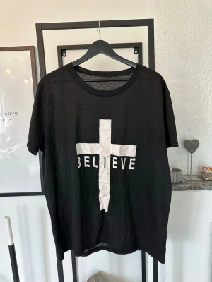 Svart t-shirt med vit kors och text - Cool svart t-shirt med ett stort vitt kors och texten 'BELIEVE' tryckt över bröstet. T-shirten har rund hals och korta ärmar, perfekt för en avslappnad och trendig stil.