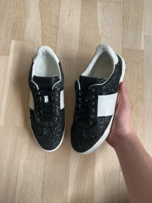 Svarta glittriga sneakers från Valentino - Snygga svarta sneakers från Valentino med glitterdetaljer och bred vit panel på sidorna. Skorna har svarta skosnören, vita sulor och insida i vitt läder. Perfekta för dig som vill sticka ut med en exklusiv touch.