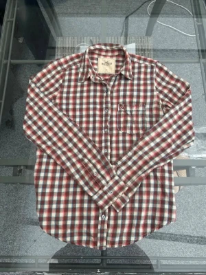 Rutig skjorta från Hollister - Säljer en långärmad rutig skjorta från Hollister i rött, vitt och blått. Skjortan har klassisk krage, knappstängning framtill och en bröstficka med broderad Hollister-logga. Perfekt för dig som gillar avslappnad och tidlös stil.