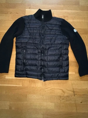 Moncler cardigan jacka  - Snygg svart dunjacka från Moncler med quiltad front och stickade ärmar. Jackan har hög krage, dragkedja framtill och två fickor med dragkedja. Moncler-logga på vänster ärm. Perfekt för kalla vinterdagar.