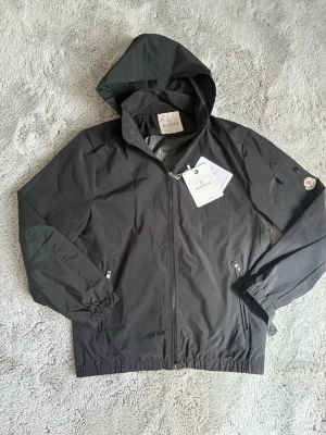 Svart vindjacka från Moncler - Snygg svart vindjacka från Moncler med huva och dragkedja framtill. Jackan har två fickor med dragkedja, elastiska muddar vid ärmar och nederkant samt Moncler-logga på ärmen. Tillverkad i lätt och slitstarkt material, perfekt för blåsiga dagar.