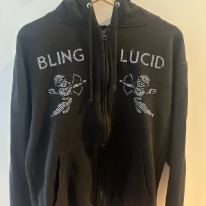 Svart hoodie BLING LUCID med strass - Svart hoodie med dragkedja från BLING LUCID. Framsidan har strassdetaljer med texten 'BLING LUCID' och två änglar. Utrustad med huva och kängurufickor. Perfekt för dig som gillar streetwear och vill sticka ut med bling.