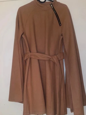 Camel cape-kappa med bälte - Stilren camel-färgad cape-kappa med bälte i midjan och vida ärmar. Hög krage med snedställd dragkedja som detalj. Kappan har en mjuk, filtliknande yta och ger en trendig siluett. Perfekt för dig som vill ha en modern och chic look.