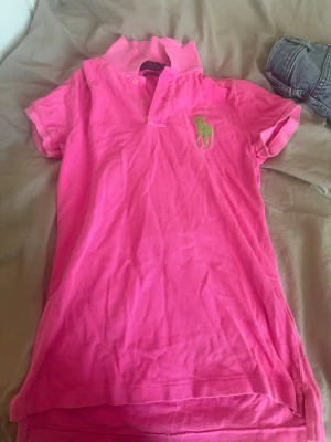 Rosa pikétröja från Ralph Lauren - Säljer en snygg rosa pikétröja från Ralph Lauren, modell The Skinny Polo. Tröjan har klassisk krage, korta ärmar och en grön broderad logga på bröstet. Perfekt för dig som gillar en sportig och färgstark stil.
