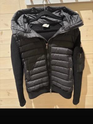 Svart cardigan från Moncler - Snygg svart cardigan från Moncler med stickade ärmar och huva. Jackan har quiltad front, två fickor med tryckknappar och dragkedja framtill. Perfekt för kalla dagar och ger en sportig men stilren look.
