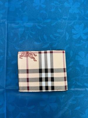 Rutig plånbok från Burberry - Säljer en klassisk rutig plånbok från Burberry i beige, svart, vitt och vinrött. Plånboken har det ikoniska Burberry-mönstret och en hästlogga i hörnet. Tillverkad i tåligt material med stilren och tidlös design.