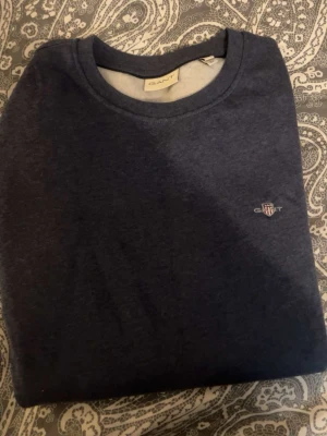 Mörkblå stickad tröja från GANT - Mörkblå stickad tröja från GANT med rund halsringning och diskret broderad logga på bröstet. Klassisk och enkel design som passar till det mesta. Tröjan är mjuk och skön, perfekt för lager på lager under kyliga dagar.
