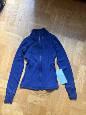 Blå Define Jacket från Lululemon - Snygg blå Define Jacket från Lululemon i mjukt och stretchigt Nulu-material. Jackan har figurnära passform, hög krage, tumhål och två dragkedjeförsedda fickor. Perfekt för träning eller som ett sportigt lager till vardags.