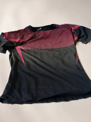 Onyx - Snygg svart t-shirt från gym shark med vinröda grafiska detaljer över bröstet och ryggen. T-shirten har korta ärmar och rund halsringning. Perfekt för dig som gillar sportig stil och vill sticka ut lite extra.