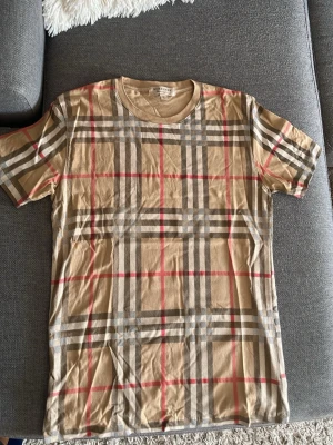 Beige rutig t-shirt från Burberry - Säljer en klassisk beige t-shirt från Burberry med det ikoniska rutiga mönstret i grått, vitt och rött. T-shirten har rund halsringning och korta ärmar. Tillverkad i mjuk bomull som känns skön mot huden. Perfekt för dig som gillar stilrena och tidlösa plagg.