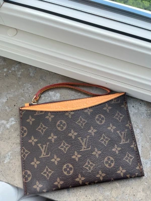 kuvertväska - Snygg kuvertväska  i klassiskt brunt monogrammönster. Väskan har guldfärgad dragkedja och handledsrem i ljusbrunt läder. Perfekt storlek för det viktigaste och en ikonisk accessoar för dig som gillar lyxig stil.