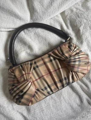Burberry rutig handväska i beige - Säljer en snygg handväska från Burberry med klassiskt beige rutmönster och mörkbrunt handtag. Väskan har unika veckade detaljer på sidorna och är tillverkad i canvas med läderdetaljer. Perfekt accessoar för dig som gillar tidlös design och lyxig känsla.