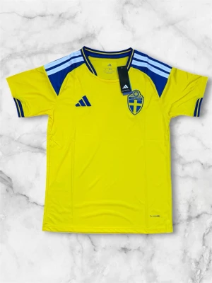 Sverige matchtröja Adidas gul/blå - Säljer en officiell Sverige matchtröja från Adidas i gult med blå detaljer och tre ränder på axlarna. Tröjan har rund hals, korta ärmar och Sveriges landslagsemblem på bröstet. Tillverkad i lätt och ventilerande polyester, perfekt för fotboll eller träning.