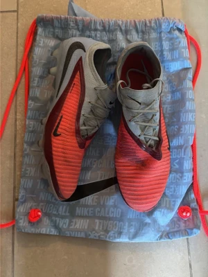 Nike röda fotbollsskor med grå detaljer - Säljer ett par snygga fotbollsskor från Nike i rött med grå snörning och detaljer. Skorna har en modern, strömlinjeformad design och är tillverkade i syntetmaterial för bra grepp och känsla på planen. Perfekta för dig som vill sticka ut på fotbollsplanen.