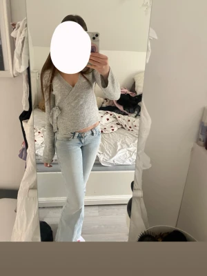 Grå omlottopp med knytband - Trendig grå omlottopp med långa ärmar och knytband i sidan. Tillverkad i mjukt stickat material som ger en avslappnad men snygg look. Perfekt att matcha med jeans för en casual stil.