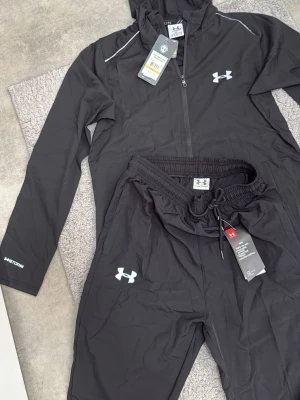 Svart träningsset från Under Armour - Säljer ett svart träningsset från Under Armour med jacka och byxor. Setet har dragkedja på jackan, huva och vita loggor på både jacka och byxa. Materialet är lätt och vattenavvisande, perfekt för löpning eller gym. Reflekterande detaljer och justerbar midja på byxorna.