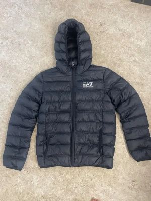 Svart dunjacka med huva från EA7 - Snygg svart dunjacka från EA7 med huva och dragkedja framtill. Jackan har ett stilrent tryck med EA7-loggan på bröstet och quiltad design som ger en modern look. Perfekt för kalla vinterdagar och enkel att matcha med olika outfits.