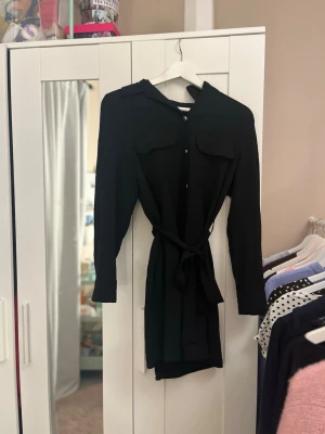 Svart skjortklänning med bälte - Stilren svart skjortklänning med långa ärmar och knytbälte i midjan. Klänningen har krage, bröstfickor och knappar framtill för en klassisk look. Perfekt för dig som gillar enkel men trendig stil.