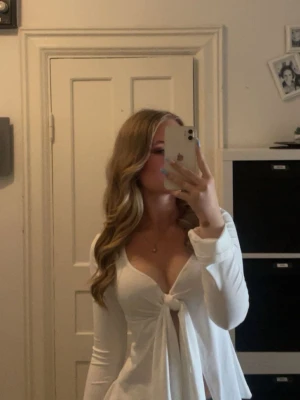 Vit omlottopp med knyt framtill - Säljer en superfin vit omlottopp med knyt framtill och djup v-ringning. Toppen har långa ärmar och är lätt utsvängd nedtill för en snygg siluett. Perfekt att styla med jeans eller kjol för en trendig look.